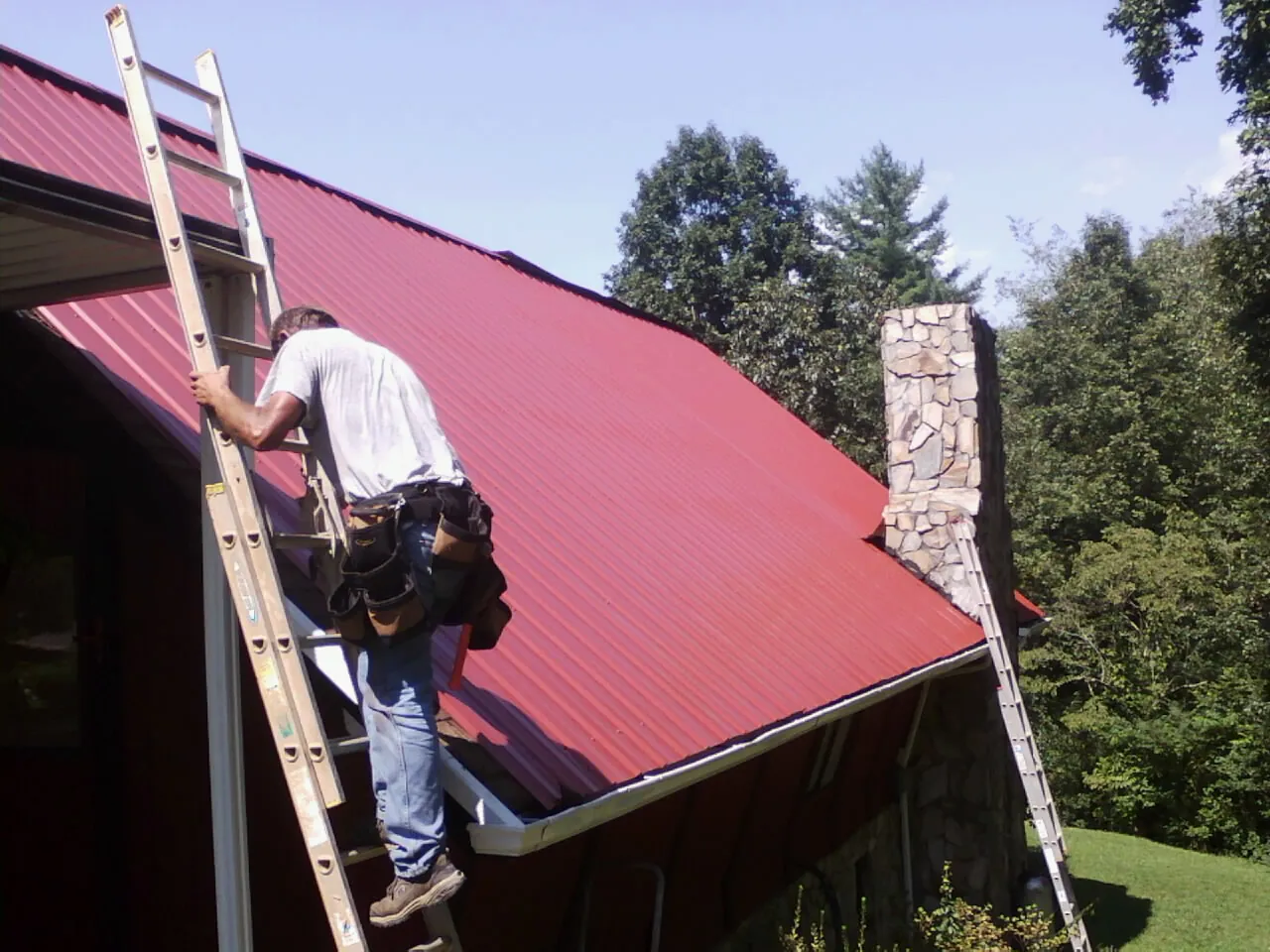 Roofer on a ladder beside a red metal roof and stone chimney — Soffit & Fascia Repair by Roofing Brothers in North Springfield